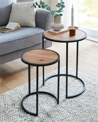 Two round side tables with wooden tops and black metal frames are placed on a textured rug next to a gray sofa. A book, a vase with greenery, and a candle are on the larger table.