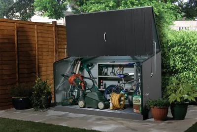 A garden shed filled with gardening tools and a bicycle, set in a backyard with a wooden fence and plants.