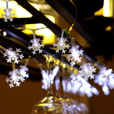 A string of snowflake-shaped LED lights hangs from a dark structure, with blurred lights and wine glasses in the background.