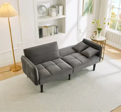 A gray sofa bed is shown in a living room setting with a floor lamp and a bookshelf in the background.
