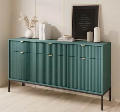 A teal sideboard with gold handles sits on black legs in a room with light wood flooring and paneled walls. Decorative items are placed on top of the sideboard.