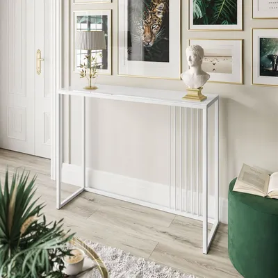 A white console table with a glass top is placed in a well-lit room, next to a white door and a collection of framed artwork on the wall. A green ottoman with a book on it is visible to the right, and a potted plant is in the foreground.