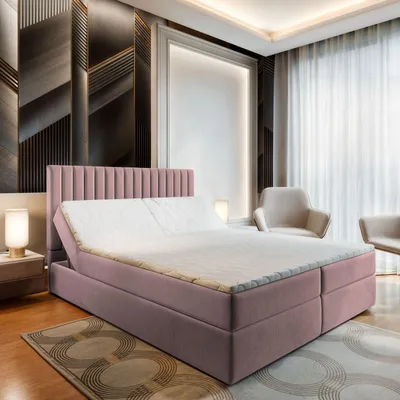 A bedroom scene featuring a pink upholstered bed with a mattress and topper, set against a decorative wall and curtains, with chairs in the background.