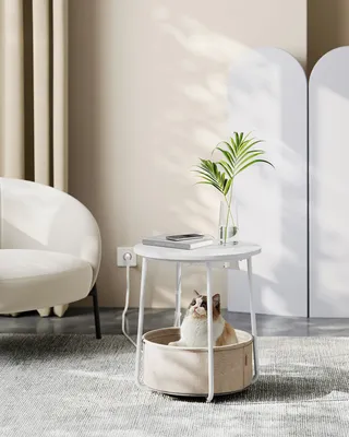 A cat sitting in a round pet bed integrated into a round side table, with a chair and curtains in the background.
