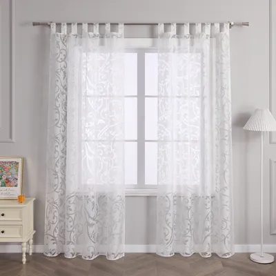 A pair of white sheer curtains with a damask pattern hanging in front of a window, with a bedside table and lamp visible.