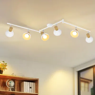 A white track light fixture with six adjustable spotlights is mounted on a white ceiling. The spotlights have white shades and wooden accents. Below the light fixture, a wooden bookshelf is visible with a globe and books.
