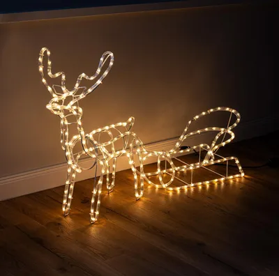 A lighted reindeer and sleigh decoration on a wooden floor against a wall.