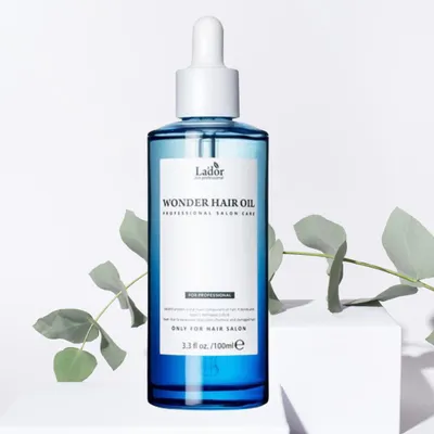 A bottle of La'dor Wonder Hair Oil is displayed on a white pedestal, with green leaves in the background.