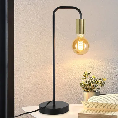 A black and gold desk lamp with a vintage bulb is placed on a white surface next to a potted plant and an open book.