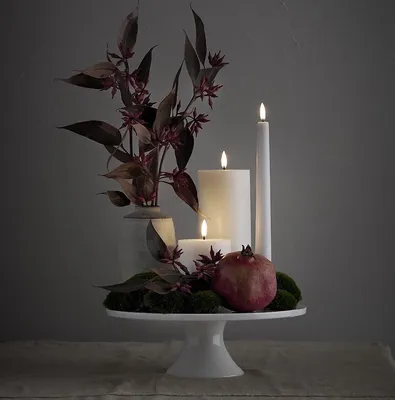 A white cake stand with LED candles, a pomegranate, moss, and decorative branches.