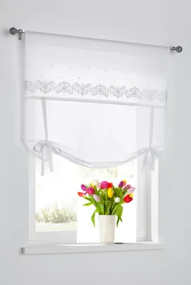A white Roman blind with decorative embroidery and tie-up ribbons is shown hanging on a window. A vase of colorful tulips sits on the windowsill below.