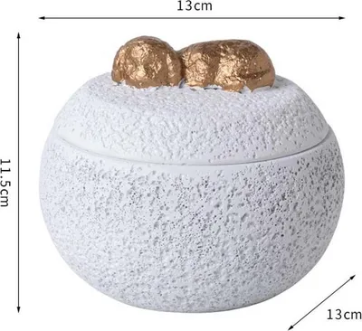 A white, textured round container with a lid, decorated with two gold-colored objects on top.