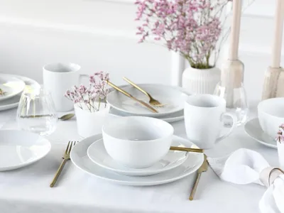 A white table setting with white porcelain dishes, gold cutlery, and a vase of pink flowers.
