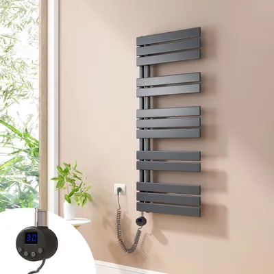 A modern anthracite electric towel radiator mounted on a light pink wall next to a window with greenery visible outside. A small white side table with a plant is also present.