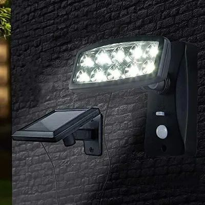 A solar-powered floodlight with a motion sensor is mounted on a dark brick wall, with its solar panel also attached to the wall.