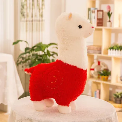 A plush toy alpaca stands on a table in a room with shelves in the background.