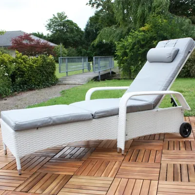 A white rattan sun lounger with gray cushions is placed on a wooden deck in a garden setting. In the background, there is a small bridge and lush greenery.