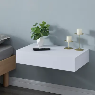 A white floating shelf mounted on a light blue wall, next to a bed. The shelf holds a plant in a vase, a remote control, and two gold candle holders with white candles.