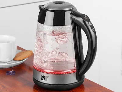 A glass electric kettle with water boiling inside, placed on a wooden table next to a cup of tea and a cookie.