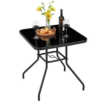 A square glass-top table with black metal legs, set up with two wine glasses, a bottle of wine, and a small woven basket with bread and flowers.