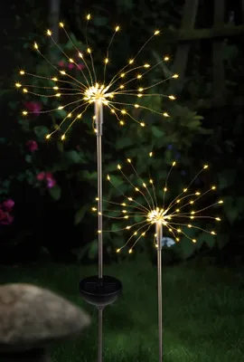 Two solar-powered firework lights are illuminated in a garden at dusk.