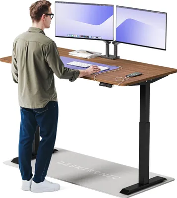 A man is standing at a height-adjustable desk with two monitors, using a mouse and keyboard. An anti-fatigue mat is on the floor.