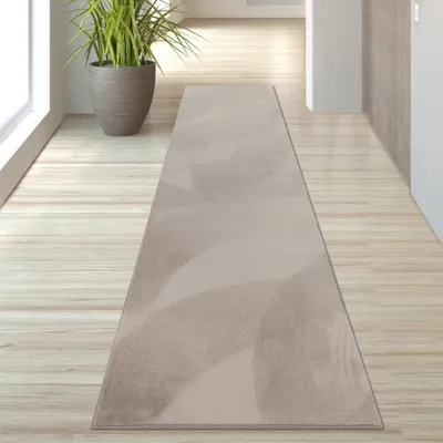 A beige runner rug with a subtle abstract pattern is laid on a light wooden floor in a hallway. A large potted plant is visible to the left.