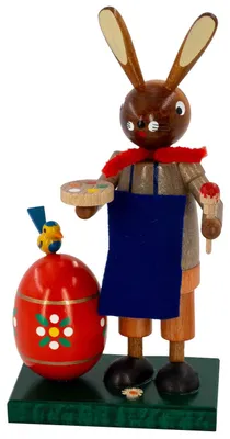Ein hölzerner Osterhase mit bemaltem Gesicht hält einen Pinsel und eine Palette in den Händen während er neben einem großen roten Osterei mit Blumenmuster steht und ein kleines gelbes Küken auf dem Ei sitzt