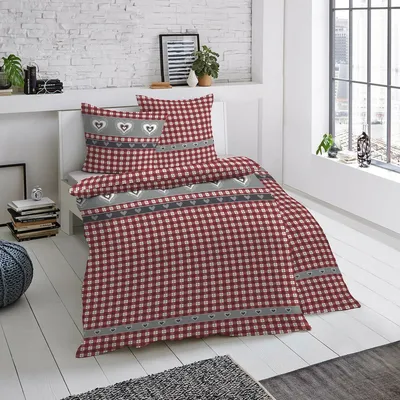 A bedroom scene with a bed made up with red and white checkered bedding featuring heart patterns, set against a white brick wall and a window.