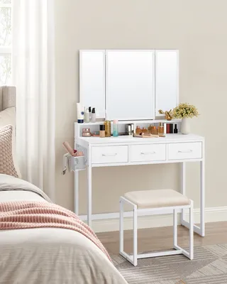 A white vanity table with a tri-fold mirror and stool, set up in a bedroom next to a bed.