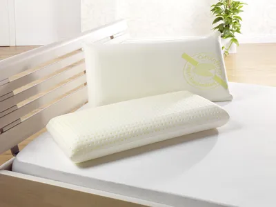 Two pillows are placed on a bed, with a wooden bed frame visible on the left and a plant in the background.