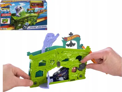 A Hot Wheels City Ghost Garage playset is shown with two hands interacting with it. A box for the playset is visible in the upper left corner.