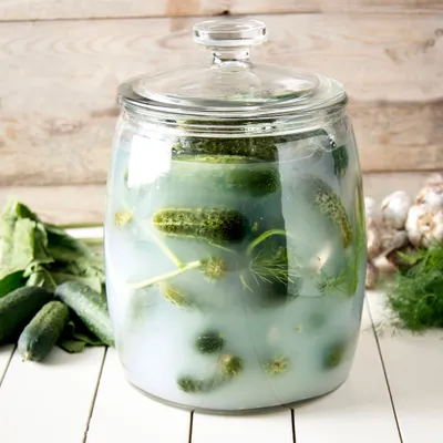 A glass jar filled with pickles and brine, surrounded by fresh cucumbers, dill, and garlic on a wooden surface.