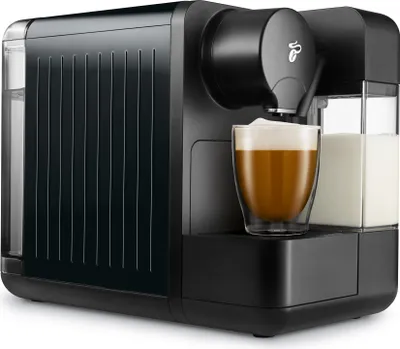 A black Tchibo Cafissimo coffee machine is shown on a white background, with a glass of coffee with foam in front of it and a container of milk to the side.