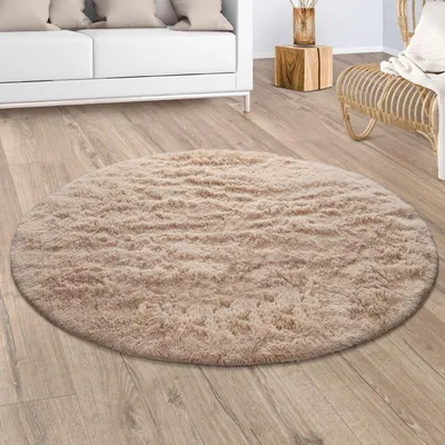A round, fluffy beige rug is placed on a wooden floor in a living room setting, with a white sofa and a wicker chair visible in the background.