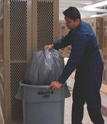 A man is putting a large black trash bag into a gray trash can next to lockers.
