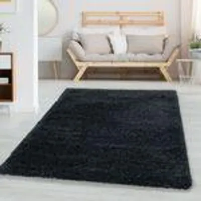 A dark, plush rug is laid out on a light-colored floor in a modern living room setting. A light-colored sofa with cushions is visible in the background.