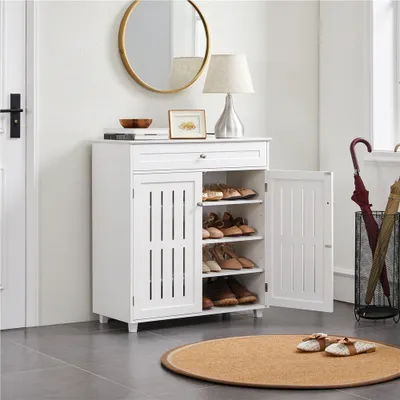 A white shoe cabinet with a drawer and two doors, one of which is open, displaying shoes on shelves. A round gold mirror and a lamp are on top of the cabinet. An umbrella stand with umbrellas is to the right.