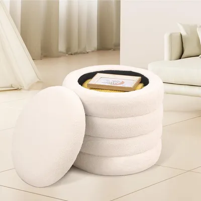 A white, fluffy storage ottoman with its lid open, revealing a framed picture and a yellow blanket inside. The ottoman is placed on a tiled floor in a well-lit room with sheer curtains in the background.