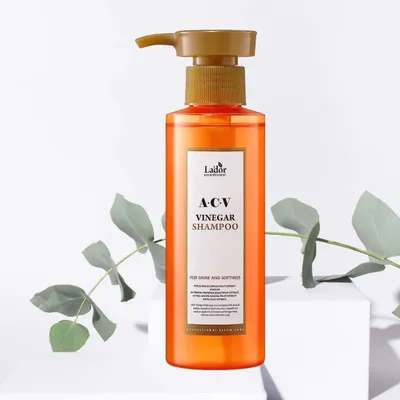 A bottle of Lador ACV Vinegar Shampoo is placed on a white pedestal, with green leaves on either side.