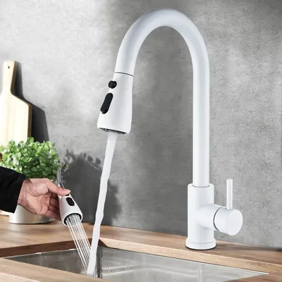A white kitchen faucet with a pull-down sprayer is shown in use over a stainless steel sink. A hand is holding the sprayer head, demonstrating its functionality.