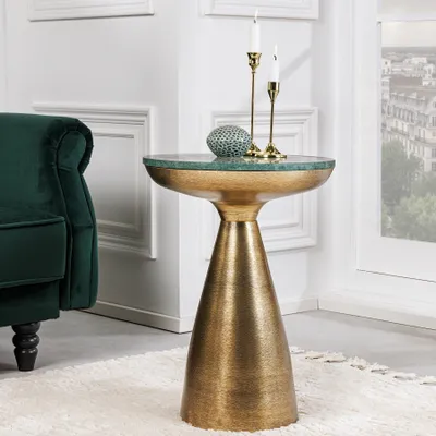 A gold accent table with a green marble top is placed next to a green velvet chair, in a room with white paneled walls and a window overlooking a city.