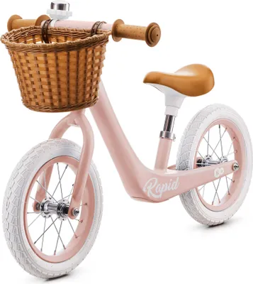 A pink balance bike with a wicker basket and brown handlebars and seat, shown against a white background.