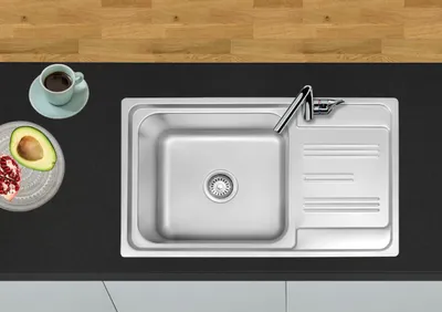 A top-down view of a stainless steel kitchen sink with a faucet, a small dish with avocado and pomegranate seeds, and a cup of coffee on a black countertop next to a wooden surface.
