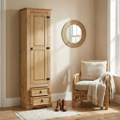 A tall wooden cabinet with two drawers at the bottom, placed next to a wicker chair with a cushion and a blanket, in a room with a round mirror on the wall.