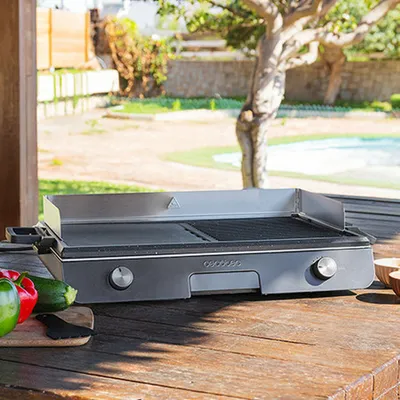 An electric grill is placed on a wooden table outdoors, with vegetables in the foreground and a pool and trees in the background.