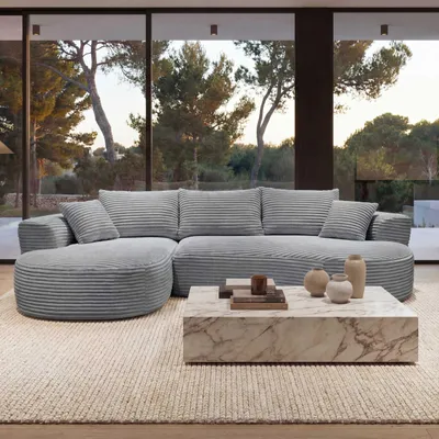 A modern living room with a large sectional sofa and a marble coffee table, with a view of trees and nature through the windows.