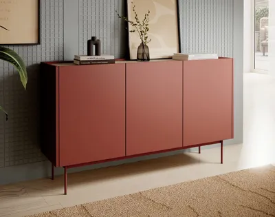 A modern red sideboard with three doors stands against a textured gray wall, adorned with framed art and decorative items on its surface. The scene is set in a well-lit room with a beige rug in the foreground.