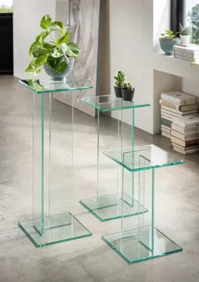 Three glass pedestal tables of varying heights are arranged on a concrete floor, with one table holding a potted plant and another holding two small potted succulents. In the background, a stack of books and a window are visible.