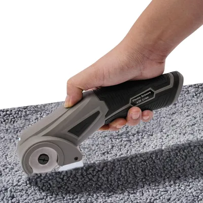 A hand is holding a cordless electric scissors and cutting a gray carpet.